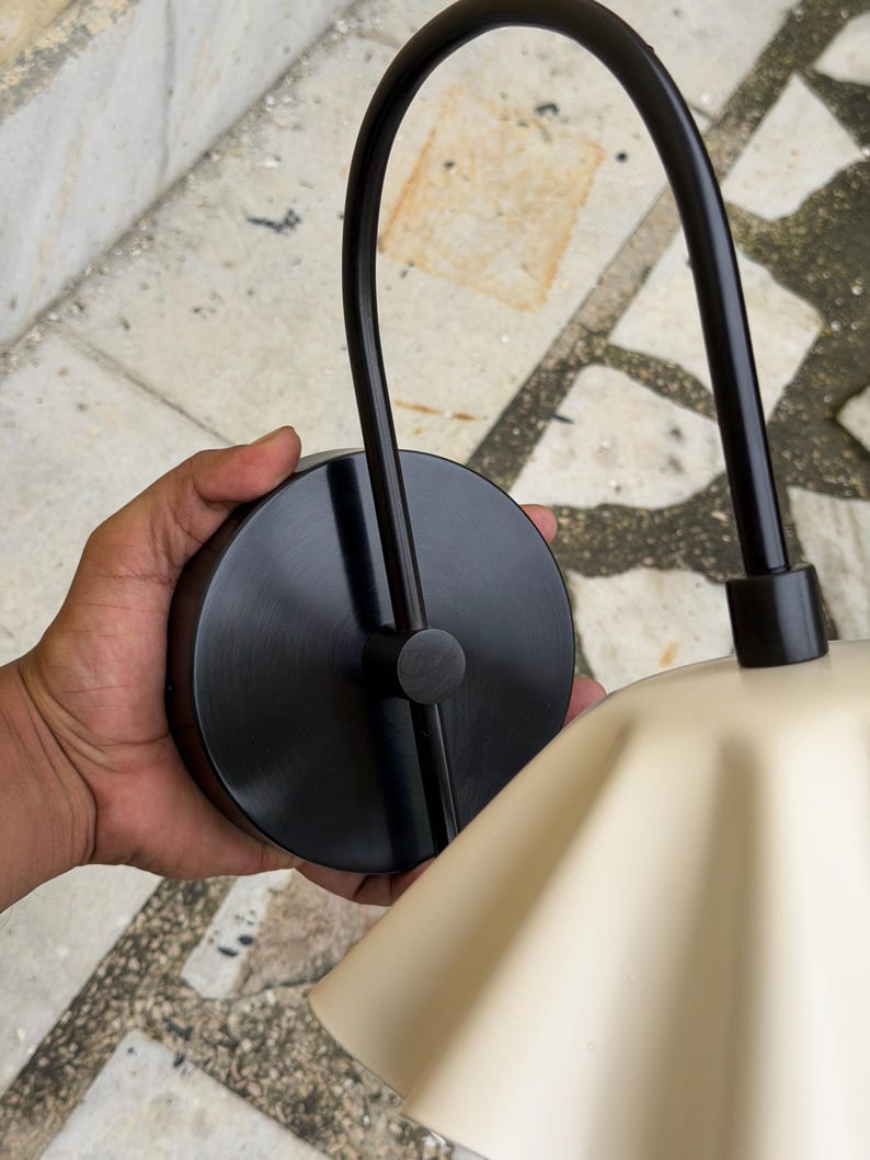 May include: A wall-mounted light fixture featuring a circular matte black base and a curved black metal arm that supports a wide, fluted, cream-colored lampshade. The design includes a central black connector piece where the arm meets the base.