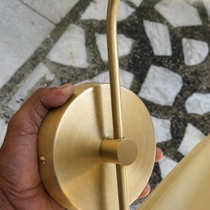 May include: A brushed brass wall light fixture featuring a circular mounting plate and a slender, curved metal arm. The finish is a matte gold tone with a subtle metallic texture. The text BRUSHED BRASS is visible in the upper right corner.