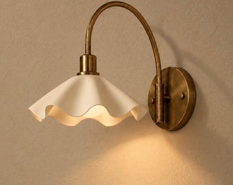 May include: A wall-mounted light fitting featuring a curved antique brass arm and a circular mounting plate. The lamp has a wide, white, ruffled-edge shade that directs light downwards, creating a soft glow on the surface below.