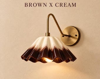 May include: A wall lamp with a brass-toned metal arm and a circular wall mount. The lamp features a fluted, bell-shaped shade with a cream-coloured top that transitions into a dark brown base. The text BROWN X CREAM is visible above.