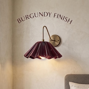 Peut inclure: Une applique murale dotée d'un abat-jour plissé bordeaux brillant et d'un bras en laiton courbé. Le texte BURGUNDY FINISH est imprimé au-dessus du luminaire. La lampe possède une plaque murale circulaire en laiton et une ampoule visible.