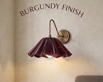 May include: A wall-mounted lamp featuring a glossy, pleated burgundy shade and a curved brass arm. The text BURGUNDY FINISH is printed above the fixture. The lamp has a circular brass wall plate and a single light bulb visible underneath.