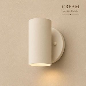 Pode incluir: Uma arandela cilíndrica com acabamento fosco na cor creme. A luminária possui uma placa de montagem circular e uma carcaça de luz vertical em formato de tubo. O texto CREAM Matte Finish está visível no canto superior direito da imagem.