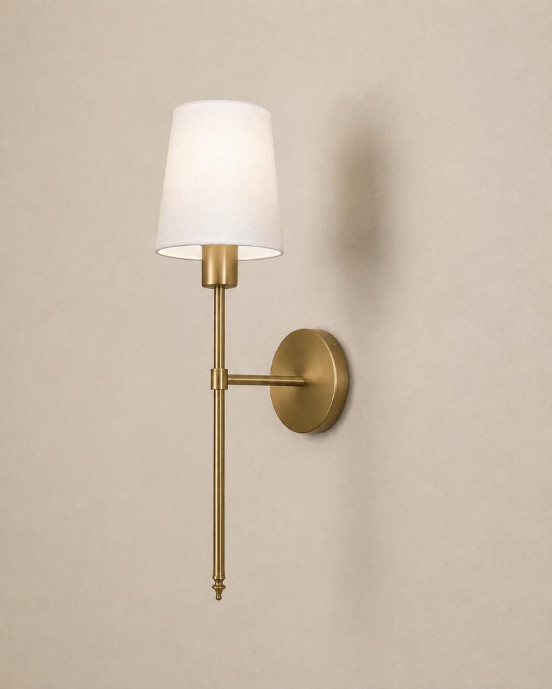 K&ouml;nnte beinhalten: Eine Wandleuchte mit einem schlanken Arm aus geb&uuml;rstetem Messing und einer runden Wandhalterung. Die Leuchte ist mit einem konischen, wei&szlig;en Stoffschirm ausgestattet, der das Licht nach unten lenkt, und besitzt ein vertikales Design mit Endst&uuml;ck.