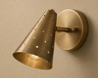 May include: A wall-mounted sconce light fitting with a brushed brass finish. The lamp features a conical shade perforated with small circular holes, attached to a circular wall plate by a short, adjustable arm with a visible hinge mechanism.