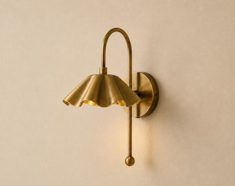 May include: A wall-mounted light fitting in a brushed brass finish. The lamp features a curved arm extending from a circular base, supporting a downward-facing, scalloped, petal-shaped shade. A straight rod extends below the shade, ending in a sphere.