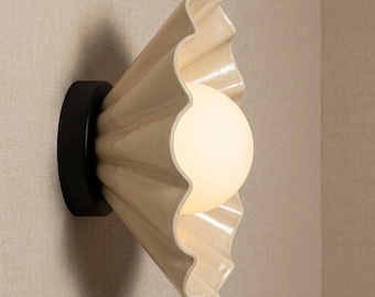 May include: A wall-mounted light fitting featuring a matte black circular base and a cream-coloured, fan-shaped ceramic shade with a ruffled edge. A spherical, frosted white glass bulb is positioned in the centre, emitting a soft, warm glow.