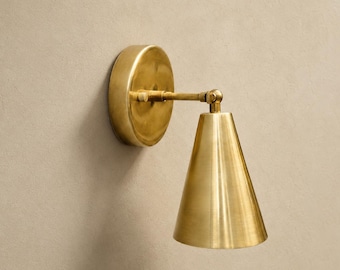May include: A wall-mounted lamp featuring a circular brass backplate and a conical brass shade. The fixture includes an adjustable arm with a pivot joint, connecting the base to the downward-facing metal cone, finished in a polished brass tone.