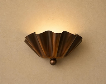 May include: A wall sconce featuring a semi-circular, pleated metal shade with a dark bronze finish. The fixture emits a warm, soft glow from the top, and a small, spherical metal knob is attached to the bottom center of the pleated structure.