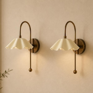 May include: A pair of wall sconces featuring a curved antique brass arm and a cream-colored, ruffled, bell-shaped shade. Each lamp is mounted on a circular brass base and includes a downward-facing light source with a small spherical finial.