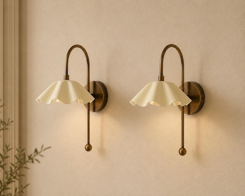 May include: A pair of wall sconces featuring a curved antique brass arm and a cream-colored, ruffled, bell-shaped shade. Each lamp is mounted on a circular brass base and includes a downward-facing light source with a small spherical finial.