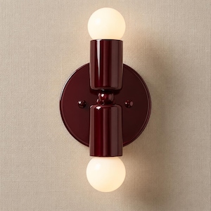 May include: A wall-mounted light fixture featuring a circular dark burgundy base and two cylindrical dark burgundy sockets. Each socket holds a round, glowing white light bulb at the top and bottom, creating a symmetrical vertical design.