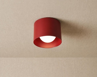 May include: A cylindrical ceiling light fixture with a matte red exterior finish. The fixture features a circular opening at the bottom that reveals a white, spherical glass light bulb centred within the red metal housing, providing illumination.