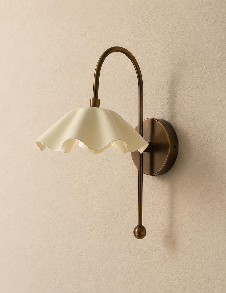 May include: A wall-mounted lamp featuring a curved, antique brass-toned metal arm and a circular wall plate. The light fixture includes a wide, off-white, scalloped-edge shade that flares outward, suspended from the top of the arched metal frame.