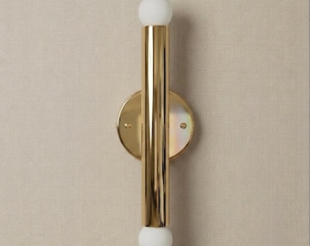 May include: A wall light featuring a vertical polished gold cylindrical body mounted on a circular gold backplate. The fixture is capped at both the top and bottom with a spherical white frosted glass globe, creating a symmetrical design.