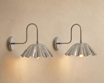 May include: A pair of wall-mounted sconces featuring a polished silver-tone finish. Each fixture has a curved arm extending from a circular base to a wide, fluted, scalloped shade. A single warm-toned light bulb is visible beneath each metal shade.