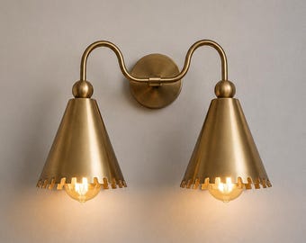 May include: A double wall sconce light fixture with a brushed brass finish. It features a circular wall mount, two curved arms, and two downward-facing conical shades with scalloped edges. Each shade houses a glowing, warm-toned light bulb.