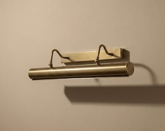 Könnte beinhalten: Eine Wandleuchte für Bilder mit gebürstetem Messing-Finish. Die Leuchte besteht aus einer rechteckigen Rückplatte, zwei geschwungenen Haltearmen und einem langen, zylindrischen Leuchtengehäuse. Das Design ist metallisch, matt und schlicht.