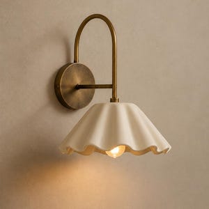 Puede incluir: Un aplique de pared con un brazo curvo de latón antiguo y una placa circular. La lámpara incluye una pantalla de cerámica de color blanco roto con bordes ondulados, orientada hacia abajo, y una bombilla que emite una luz cálida.