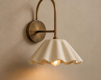 May include: A wall-mounted light fitting featuring a curved antique brass arm and a circular wall plate. The lamp includes a scalloped, off-white ceramic shade with a downward-facing opening and a single exposed light bulb emitting a warm glow.