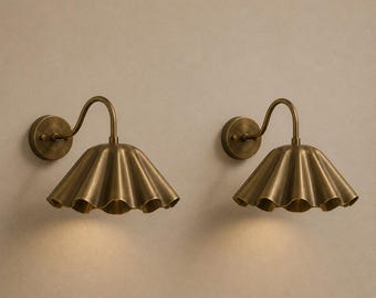 May include: A pair of wall-mounted lamps featuring a metallic brass finish. Each lamp has a circular wall plate, a curved arm, and a wide, fluted, bell-shaped shade with a scalloped edge that directs light downwards in a soft, warm glow.