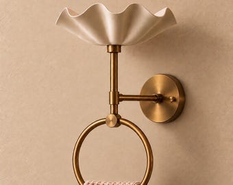 May include: A wall-mounted bathroom fitting featuring a brass-toned circular towel ring and a vertical support arm topped with a cream-coloured, scalloped-edge dish. A textured, light pink towel hangs from the bottom ring.