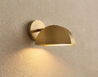 May include: A wall-mounted light fitting featuring a brushed brass finish. The lamp has a circular base plate connected to a semi-circular, dome-shaped shade that directs light downwards, creating a warm glow on the surface below the fitting.