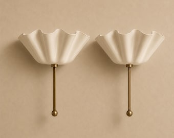 May include: A pair of wall sconces featuring off-white, ruffled, bowl-shaped shades attached to slender, vertical brass-toned metal rods. Each rod terminates in a small, spherical finial at the bottom, providing a clean and symmetrical design.