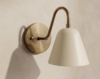 May include: A wall-mounted lamp featuring a circular, brushed brass backplate and a curved arm that supports a smooth, off-white, bell-shaped shade. The fixture has a metallic finish on the mounting hardware and a matte finish on the shade.