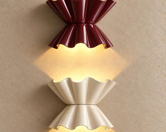 May include: Two wall sconces with a double-flared, ruffled design. The top sconce is a deep burgundy red, and the bottom sconce is a creamy off-white. Both fixtures emit a warm, glowing light from their centers, casting beams upward and downward.