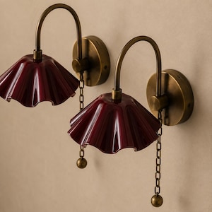 Op de afbeelding: Een paar wandlampen met gebogen messing armen en ronde messing muurplaten. Elke lamp heeft een glanzende, dieprode, geplooide glazen kap en een messing trekketting met een klein bolvormig gewichtje aan het uiteinde voor handmatige bediening.
