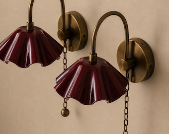 May include: A pair of wall-mounted lamps featuring curved brass arms and circular brass wall plates. Each lamp has a glossy, deep red, ruffled glass shade and a brass pull chain with a small spherical weight at the end for manual operation.