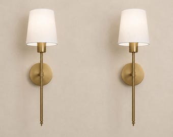 May include: A pair of wall sconces featuring a brushed brass finish, a circular wall plate, a slender vertical rod, and a white tapered fabric shade. The light fixtures are mounted vertically, providing a clean and symmetrical appearance.