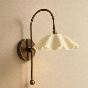 May include: A wall-mounted lamp featuring a curved antique brass arm and a circular base. The fixture includes a cream-colored, ruffled, bell-shaped shade that extends outward, complemented by a vertical brass rod ending in a small sphere.