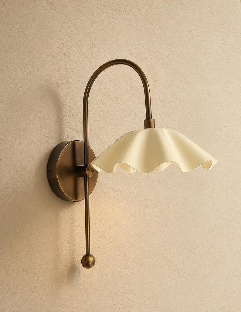 May include: A wall-mounted lamp featuring a curved antique brass arm and a circular base. The fixture includes a cream-colored, ruffled, bell-shaped shade that extends outward, complemented by a vertical brass rod ending in a small sphere.