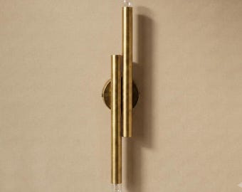 May include: A wall-mounted light fitting featuring two vertical brass-toned cylindrical tubes of unequal length. The tubes are attached to a circular brass base plate, with a small, clear light bulb visible at the top and bottom ends of the metal structure.