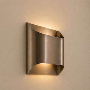 May include: A wall-mounted light fitting with a curved, metallic bronze-toned finish. The design features a rectangular shape with concave sides that allow light to emit from both the top and bottom openings, casting a warm glow on the wall.