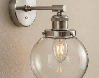 May include: A wall-mounted light fitting featuring a brushed nickel finish, a circular backplate, and a horizontal arm extending to a socket. A clear, spherical glass globe shade surrounds a single teardrop-shaped LED filament light bulb.
