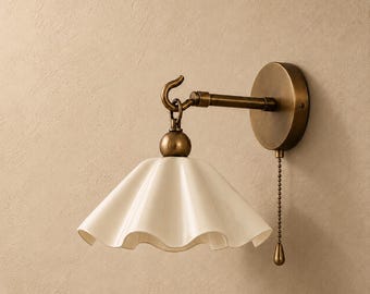 May include: A wall-mounted lamp featuring a brass-toned metal arm and a circular base. A white, ruffled, bell-shaped shade hangs from a hook on the arm. A pull chain with a small teardrop-shaped weight hangs down from the base of the lamp.
