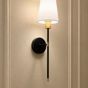 K&ouml;nnte beinhalten: Eine Wandleuchte mit einem mattschwarzen, runden Sockel und einem langen, schlanken schwarzen Stab. Die Leuchte verf&uuml;gt &uuml;ber goldfarbene Metallakzente an den Verbindungsst&uuml;cken und einen wei&szlig;en, konischen Stoffschirm, der ein warmes Licht abgibt.