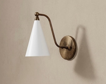 Może przedstawiać: Lampa ścienna ze stożkowym białym kloszem przymocowanym do wygiętego metalowego ramienia o wykończeniu z antycznego mosiądzu. Oprawa zawiera okrągłą mosiężną płytkę ścienną połączoną z ramieniem, które podtrzymuje skierowany w dół biały stożek.