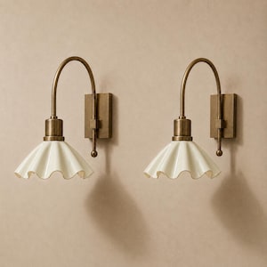 May include: A pair of wall-mounted light fixtures featuring curved antique brass arms and rectangular backplates. Each lamp has a white, scalloped, fluted ceramic shade that flares outward, suspended from a brass socket and a vertical rod.