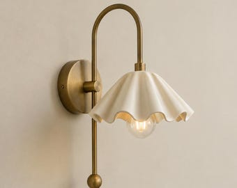 Könnte beinhalten: Eine Wandleuchte mit einem geschwungenen Messingarm und einer runden Wandplatte. Die Lampe verfügt über einen cremefarbenen, gewellten Keramikschirm mit gerüschtem Rand und einer einzelnen, nach unten gerichteten Glühbirne in der Mitte.
