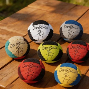 May include: A collection of seven colorful Dirtbag balls, each with a unique color scheme. The balls feature a black and white design, with the text "Dirtbag" and a stick figure logo. The balls are arranged on a wooden surface.