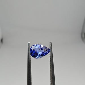 Op de afbeelding: Een peervormige, gefacetteerde edelsteen met een diepe violetblauwe kleur, vastgehouden door een metalen pincet. De steen vertoont heldere interne reflecties en een gepolijst oppervlak, wat de precieze slijpvorm en levendige kleur benadrukt.