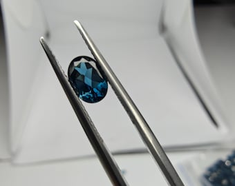Op de afbeelding: Een ovaal geslepen London blue topaas edelsteen met een gefacetteerd oppervlak, vastgehouden door een zilverkleurige metalen pincet. De edelsteen heeft een diepe, verzadigde blauwe kleur en reflecteert licht door de precieze geometrische slijpvorm.