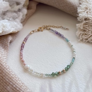 May include: A delicate beaded bracelet featuring a gradient of small faceted gemstones in shades of pink, purple, blue, and green, interspersed with white pearls and small gold-toned metal beads, finished with a gold-toned metal clasp and extender chain.