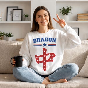 May include: A white long-sleeve sweatshirt featuring the word DRAGON in blue letters above a blue star and red and blue horizontal lines. Below this, a large red Chinese character is filled with small white stars on the front of the garment.