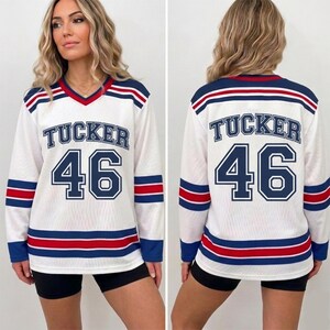 May include: A white long-sleeve jersey with a V-neck collar, featuring the name TUCKER and the number 46 in navy blue with white outlines. The sleeves and hem have horizontal stripes in navy blue and red. The fabric has a mesh texture.