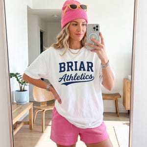 May include: A white short-sleeved t-shirt with the text BRIAR Athletics printed in dark blue, paired with pink shorts and a matching pink beanie. The t-shirt features a block letter font for BRIAR and a cursive script font for Athletics.